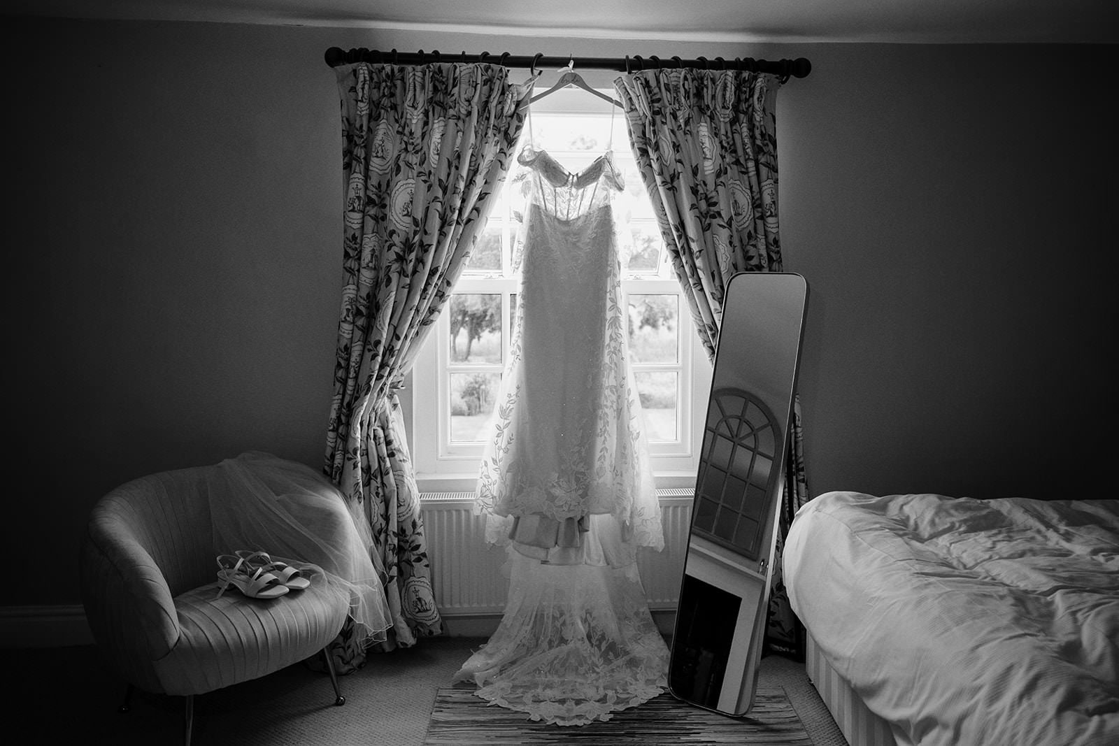 Black and white photo of a lace wedding dress hanging in a bedroom window beside a mirror and bridal shoes