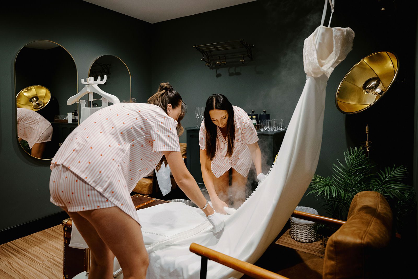 Two bridesmaids in matching pyjamas steaming the wedding dress in a dark green dressing room