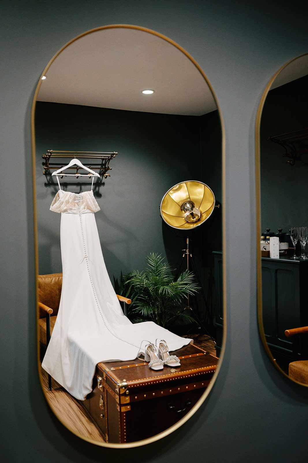 Wedding dress hanging above a leather trunk with bridal shoes, reflected in a gold oval mirror