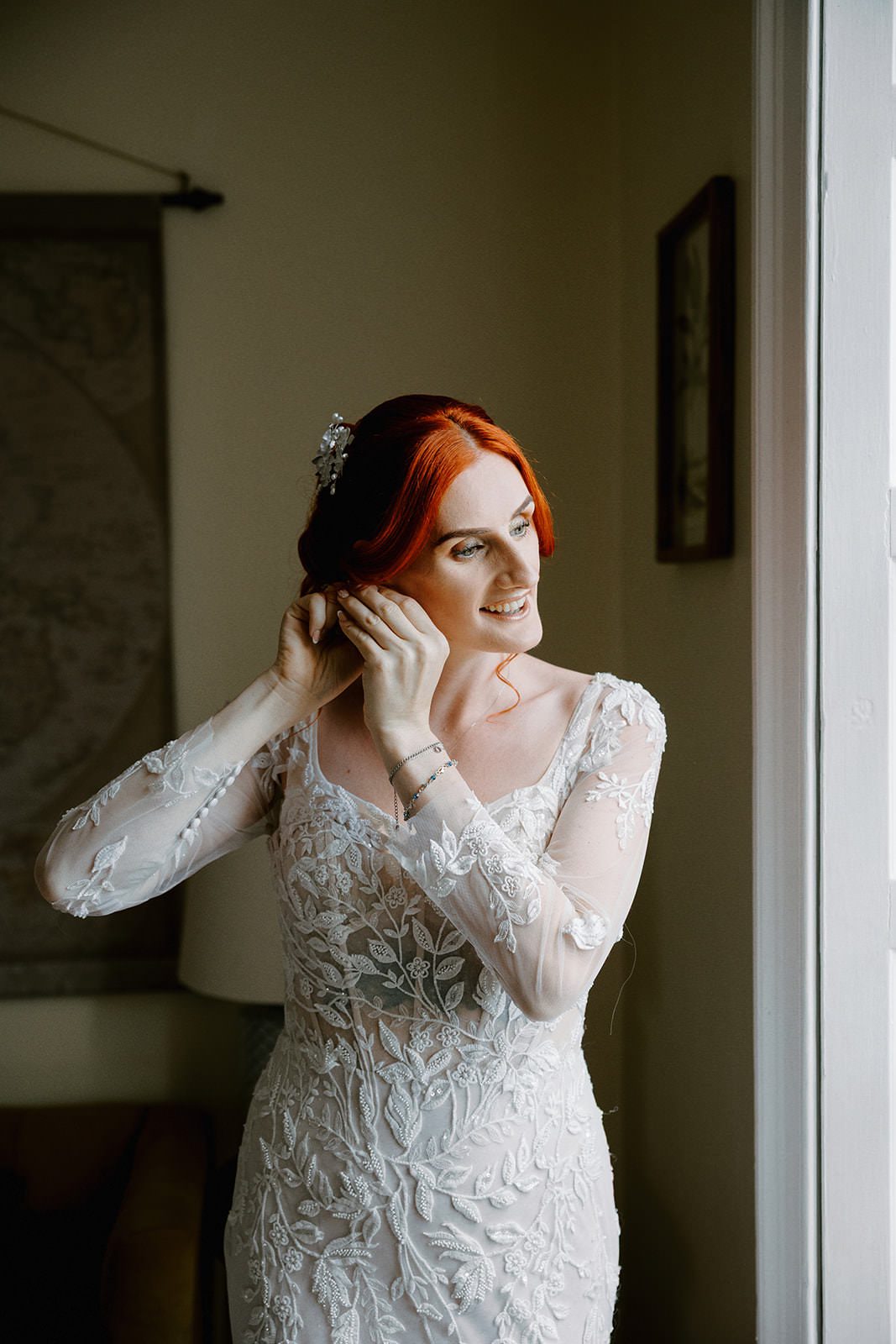 Bride with copper hair and a pearl headpiece putting on her earrings by the window