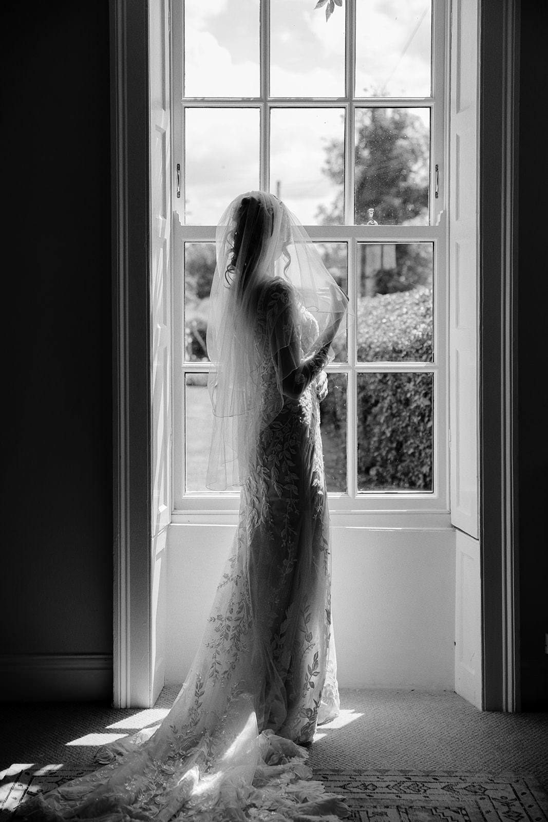Black and white photo of the bride in her veil silhouetted against a tall sash window