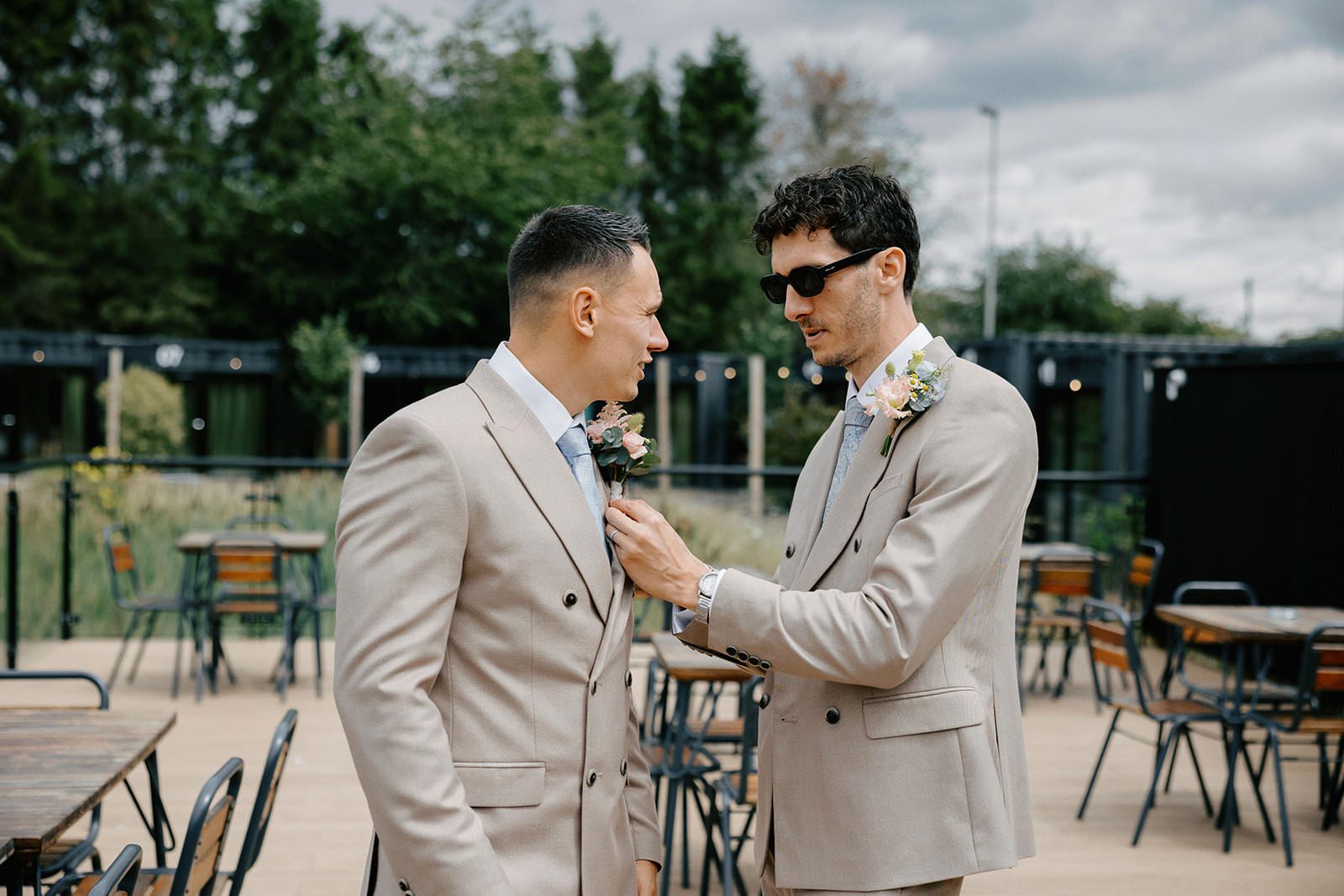 Groomsman in sunglasses pinning a floral buttonhole onto the groom's beige double-breasted suit