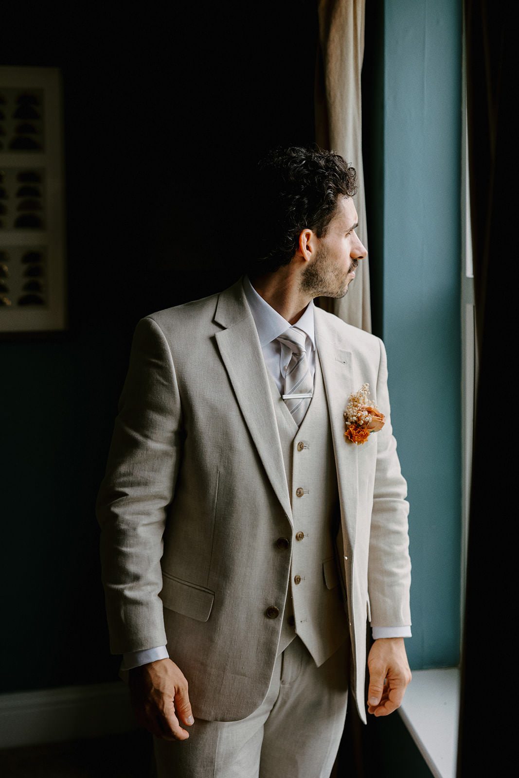 Groom in a light stone three-piece suit with a dried flower buttonhole looking out of a window