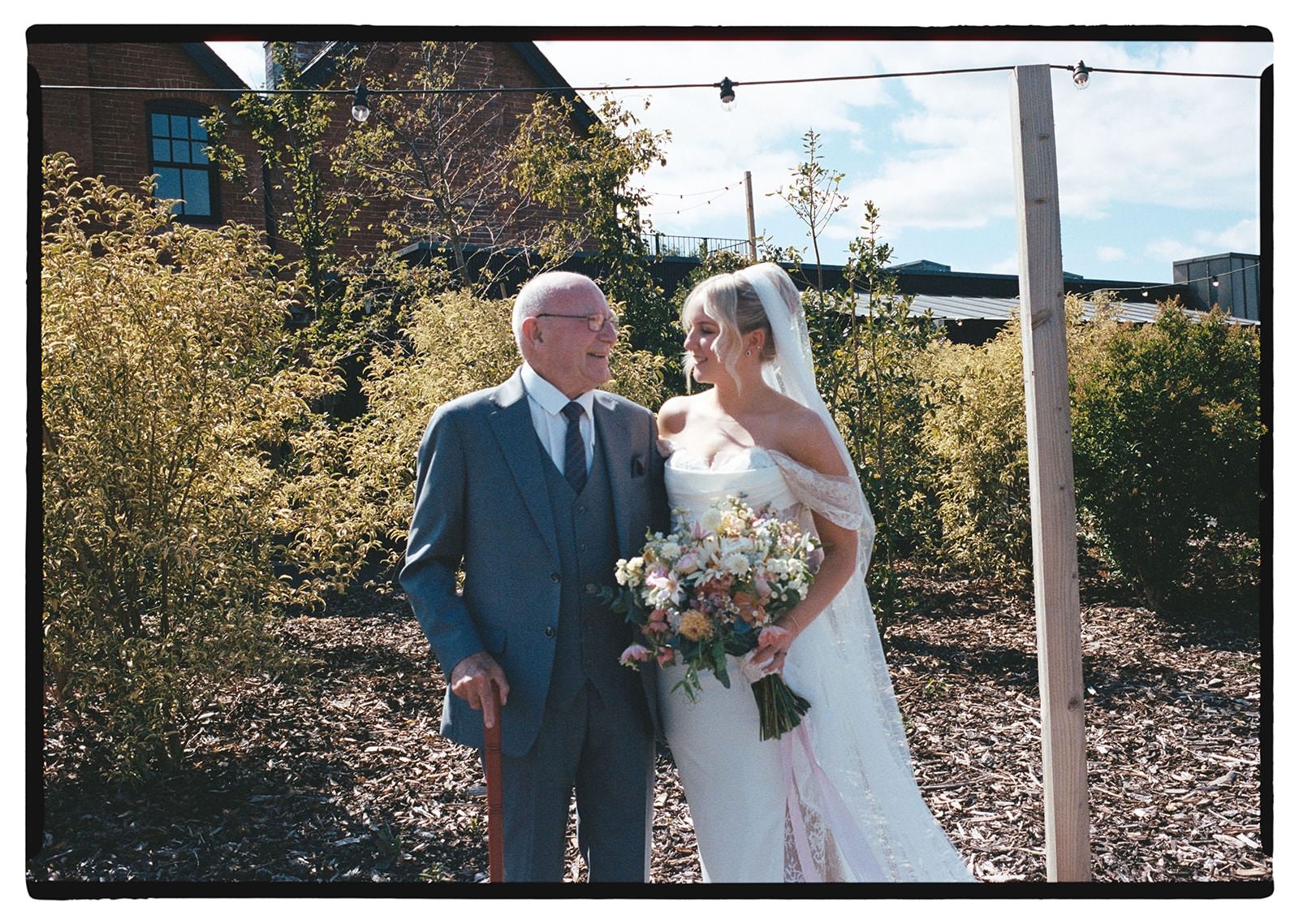 35mm film photo of the bride holding her wildflower bouquet beside her grandad in the Whistle Barns gardens
