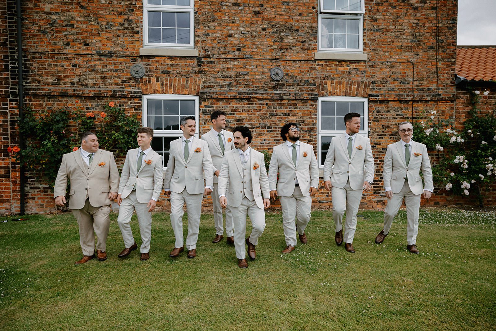 Groom and groomsmen in stone suits walking across a lawn in front of a red-brick farmhouse with roses