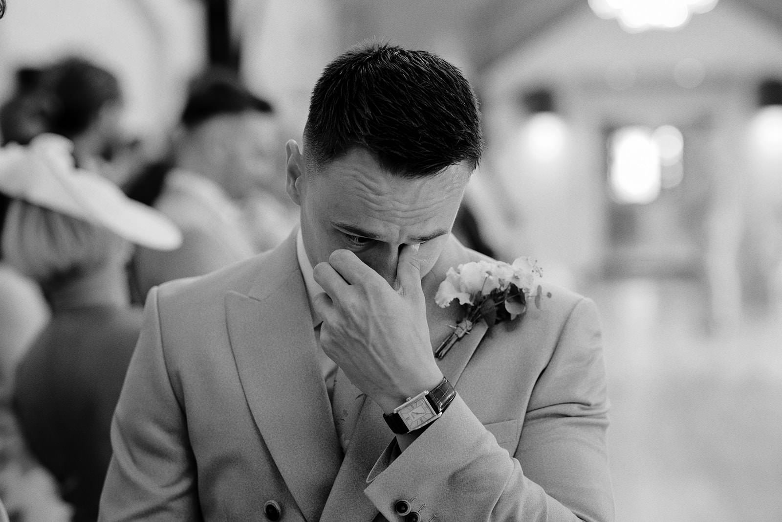 Black and white photo of the groom wiping away tears as his bride walks down the aisle