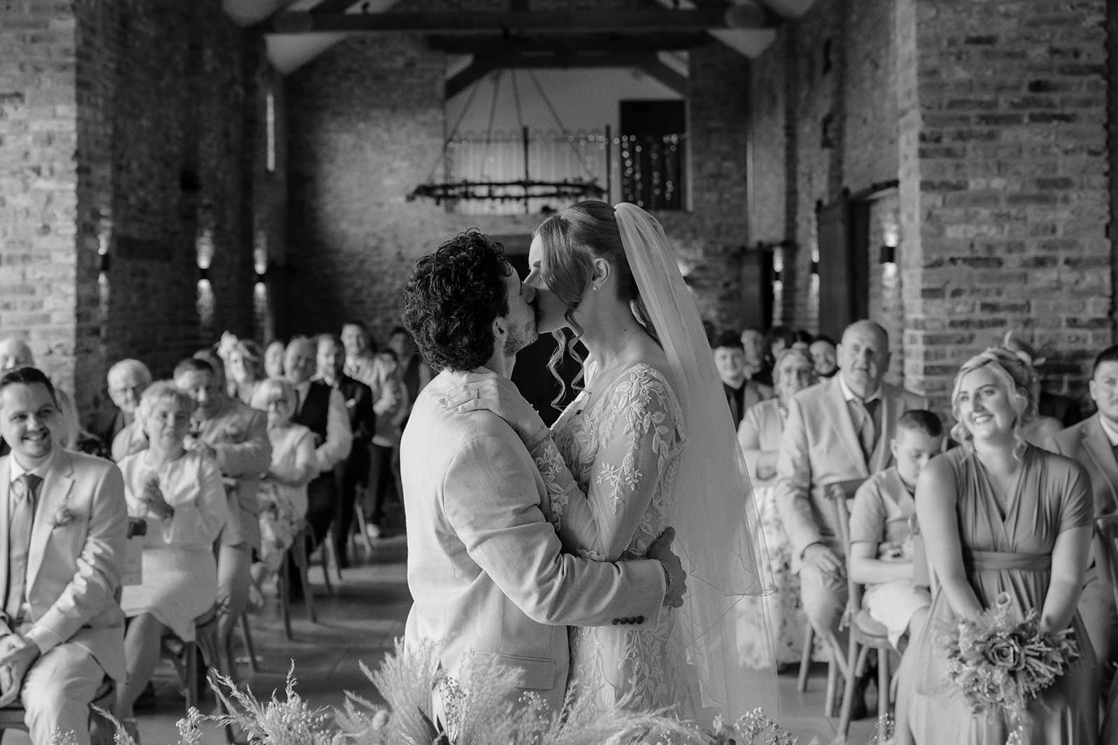 Black and white photo of the bride and groom's first kiss in front of their guests in the ceremony barn
