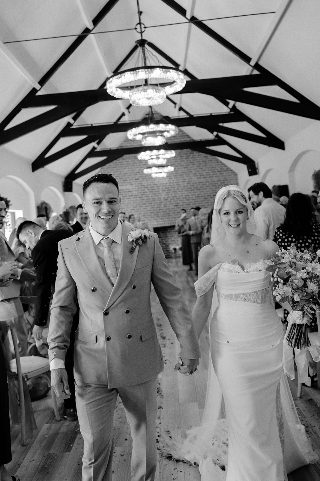 Black and white photo of the newlyweds smiling and holding hands as they walk back down the aisle