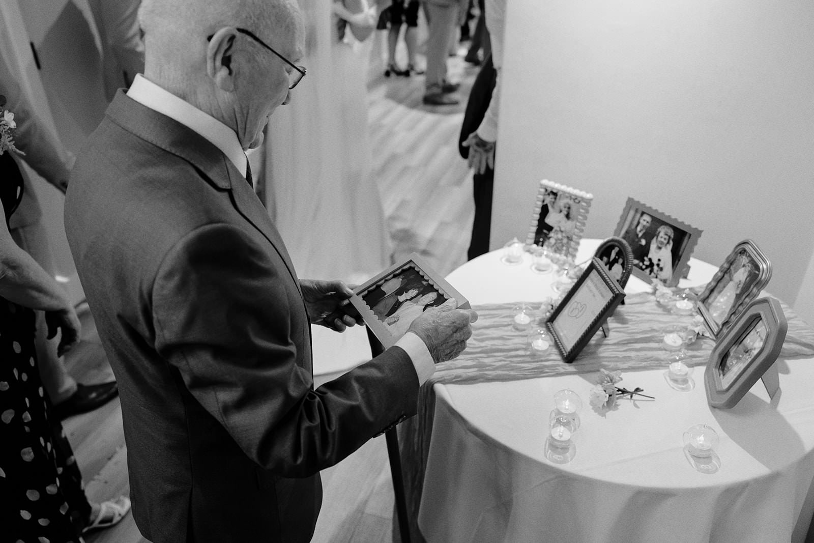 Black and white photo of the bride's grandad looking at framed family wedding photographs on a table