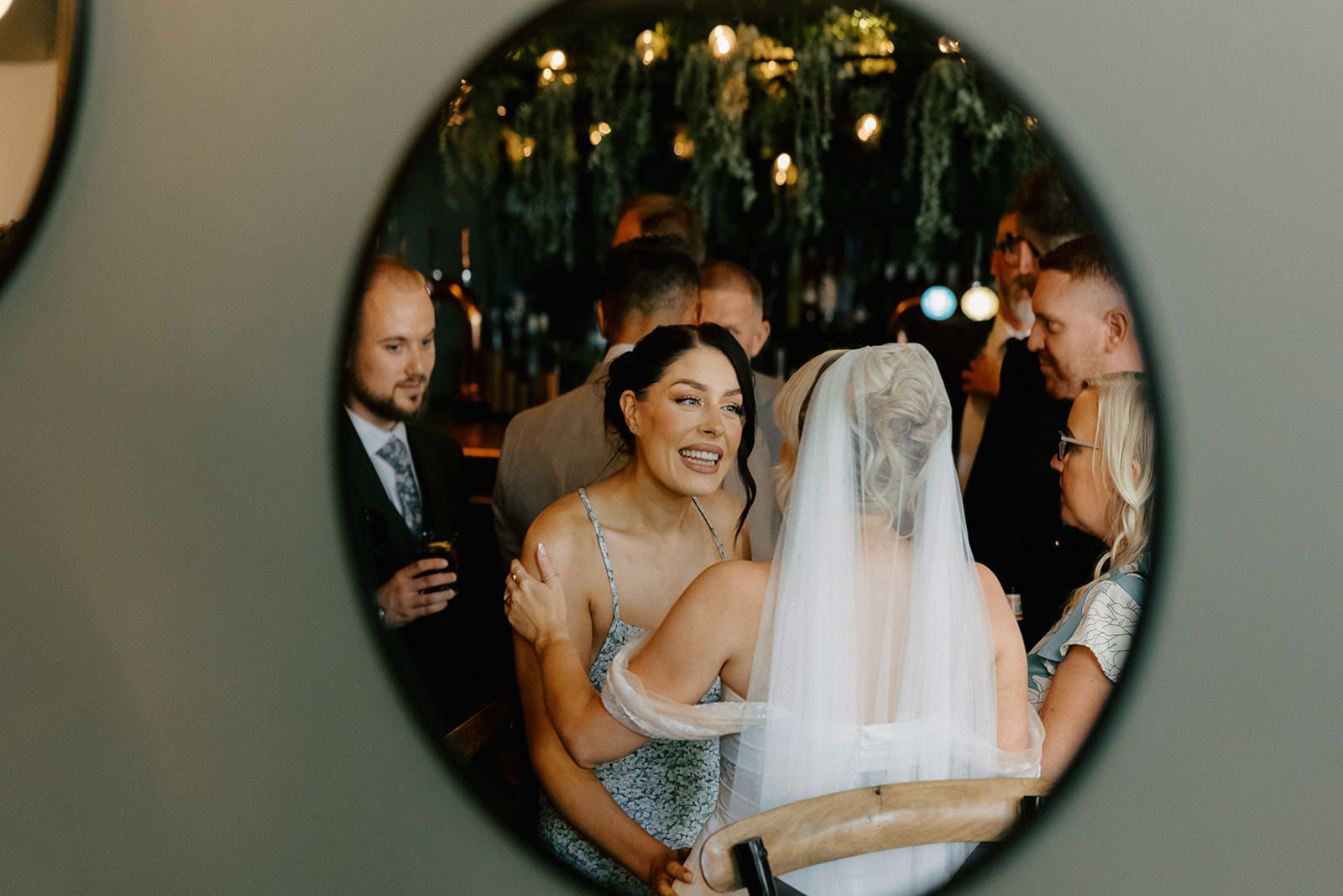 Bride hugging a guest, framed in a round mirror at the drinks reception