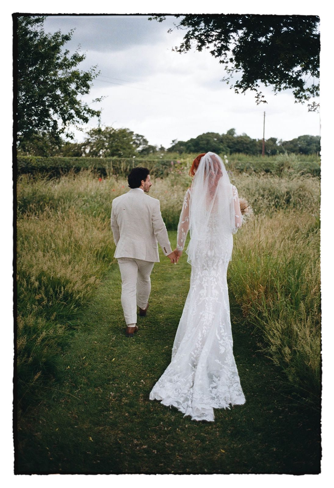 35mm film photo of the bride and groom walking hand in hand along a mown path through a wild meadow