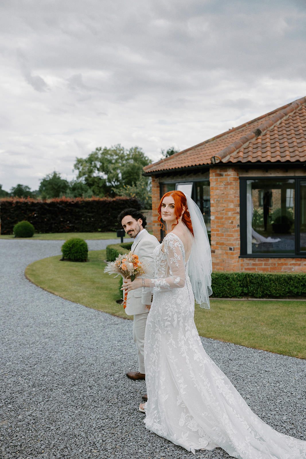 Bride looking back over her shoulder with her bouquet as the groom stands beside her on the gravel driveway