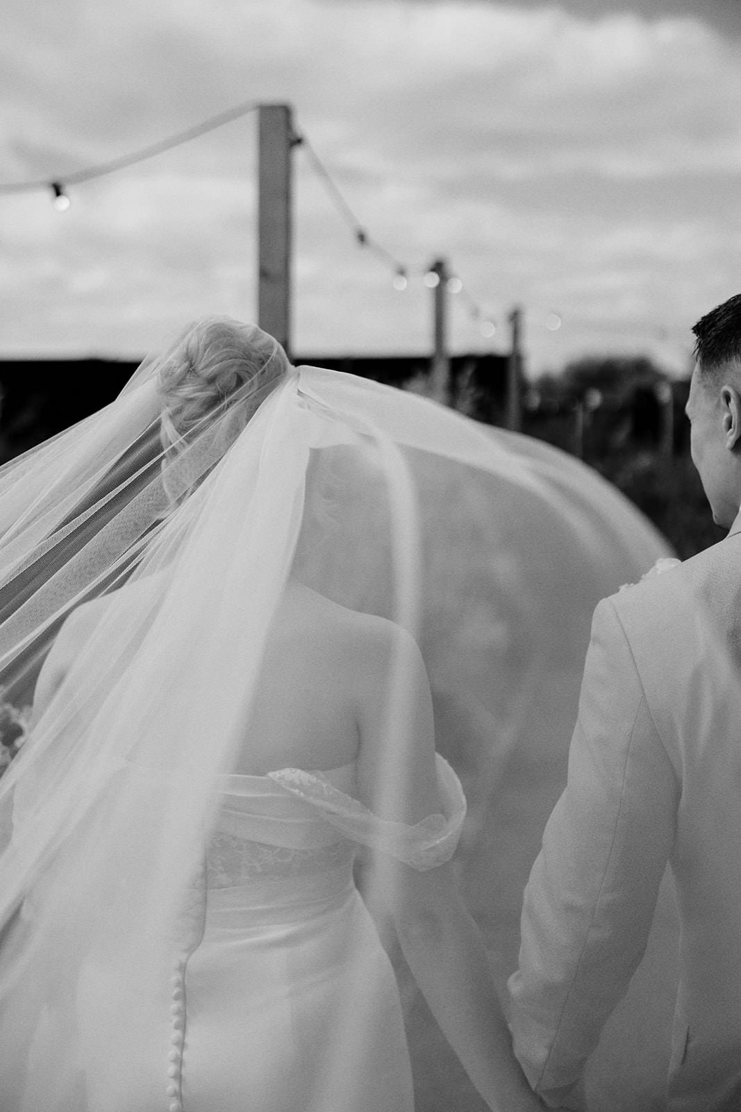 Black and white photo of the bride's long veil blowing in the wind as she walks with the groom