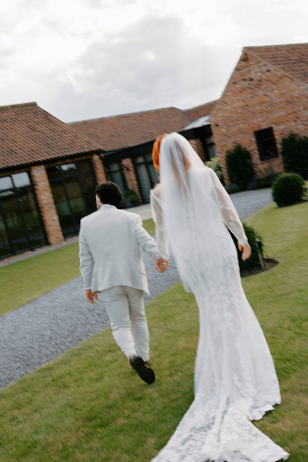 Bride and groom walking hand in hand across the lawn towards the red-brick barns, her train behind her