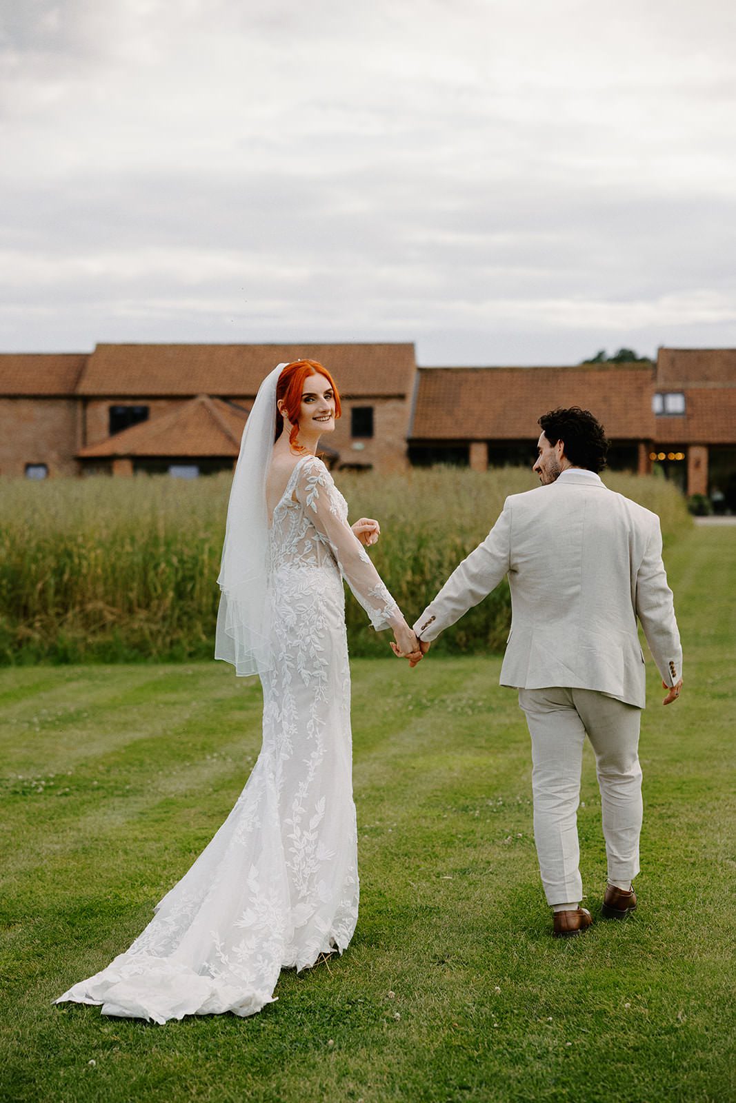 Bride smiling over her shoulder as she and the groom walk hand in hand across a striped lawn