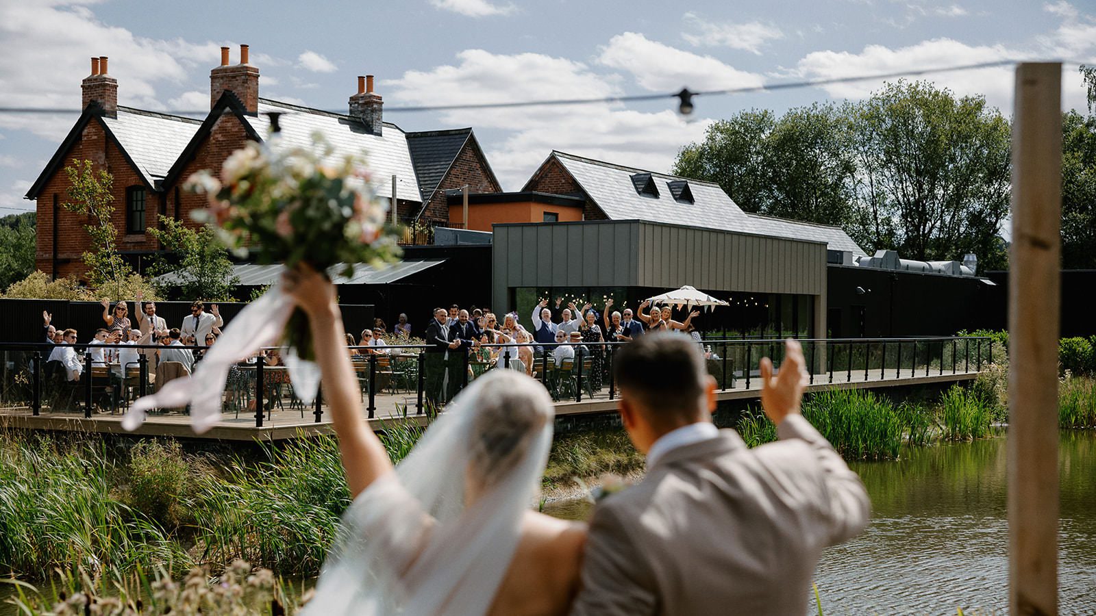 Bride raising her bouquet and groom waving to guests on the lakeside terrace at Whistle Barns