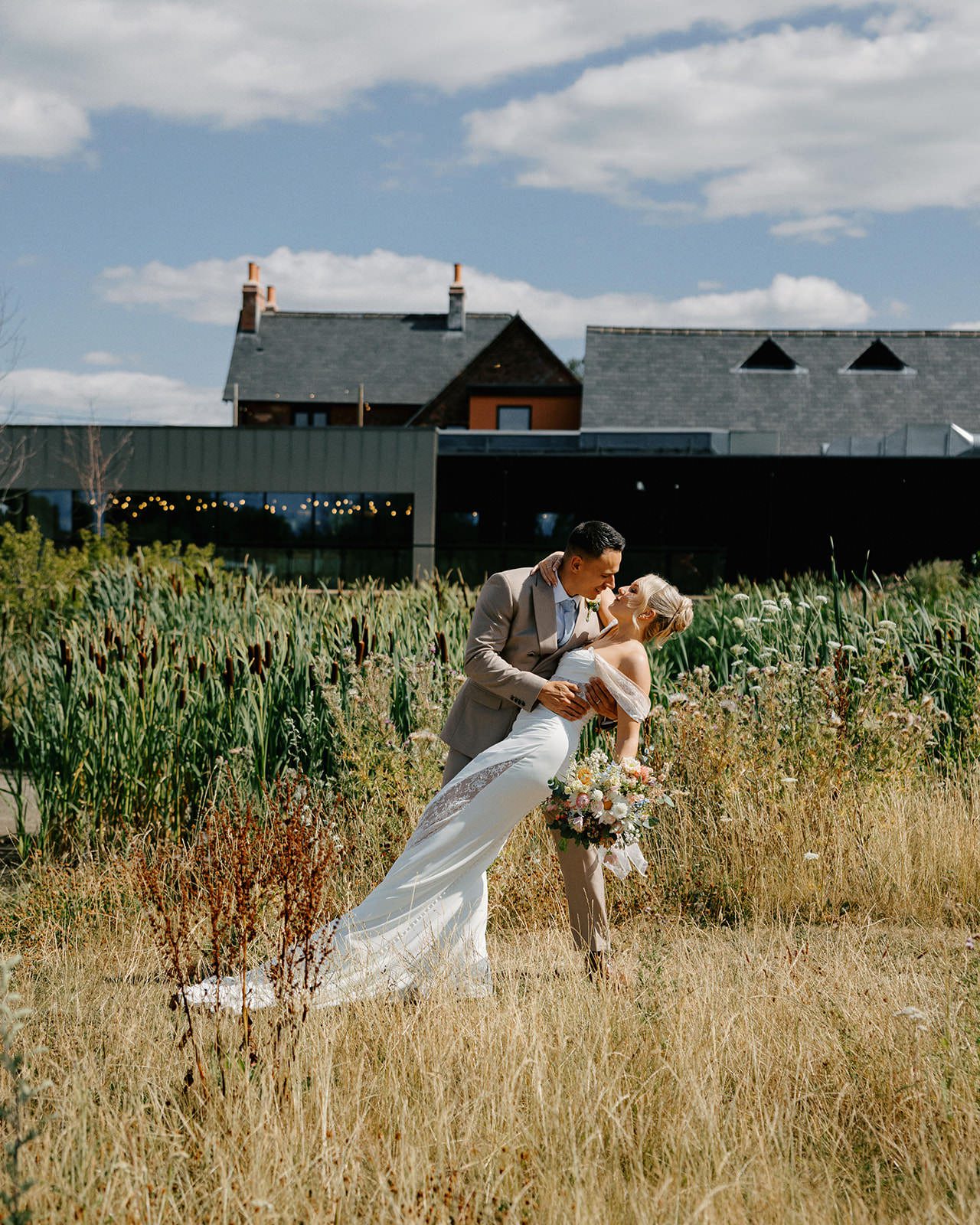 Groom dipping the bride for a kiss in long summer grass with the Whistle Barns buildings behind