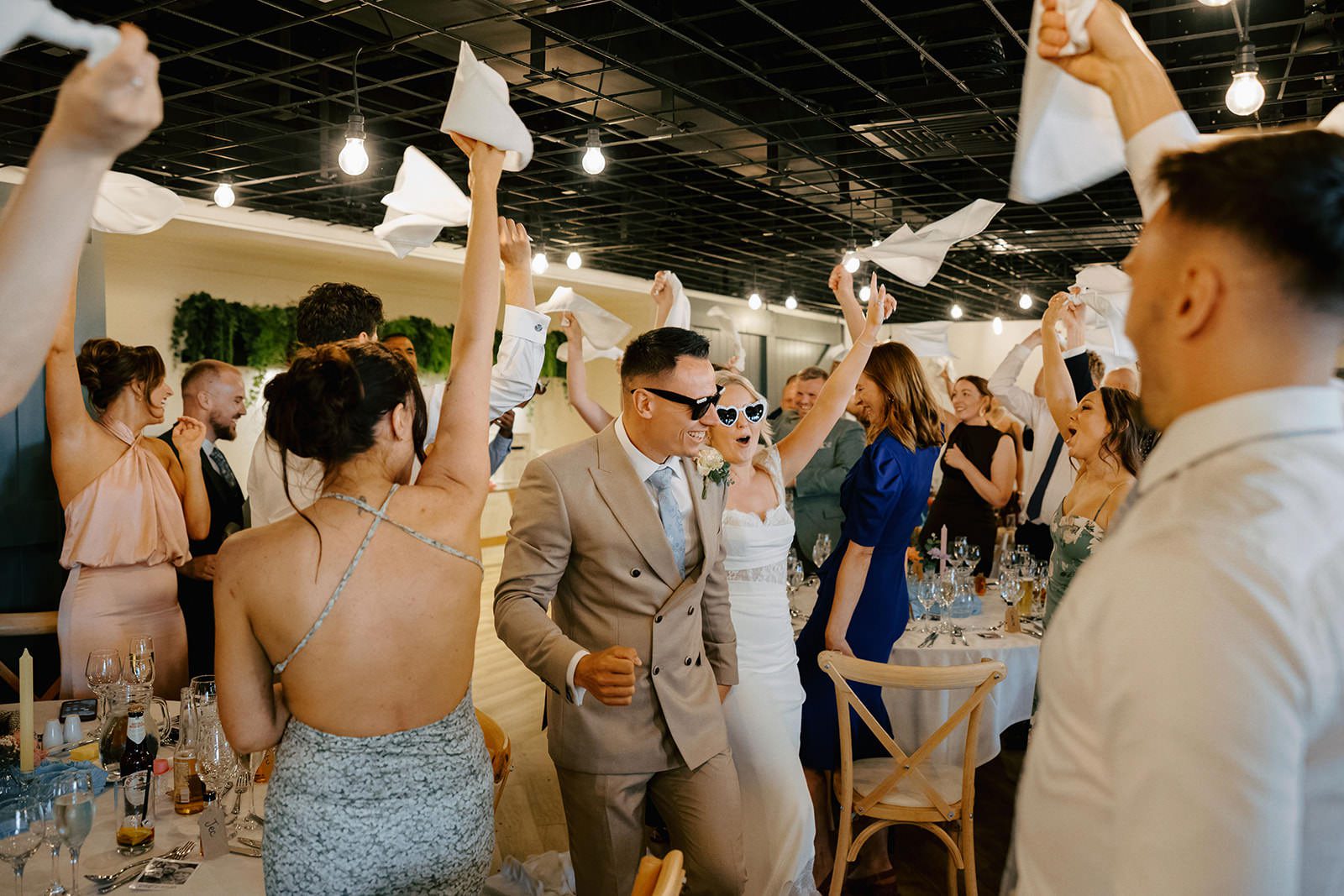 Guests waving white napkins as the bride and groom in heart sunglasses enter the wedding breakfast