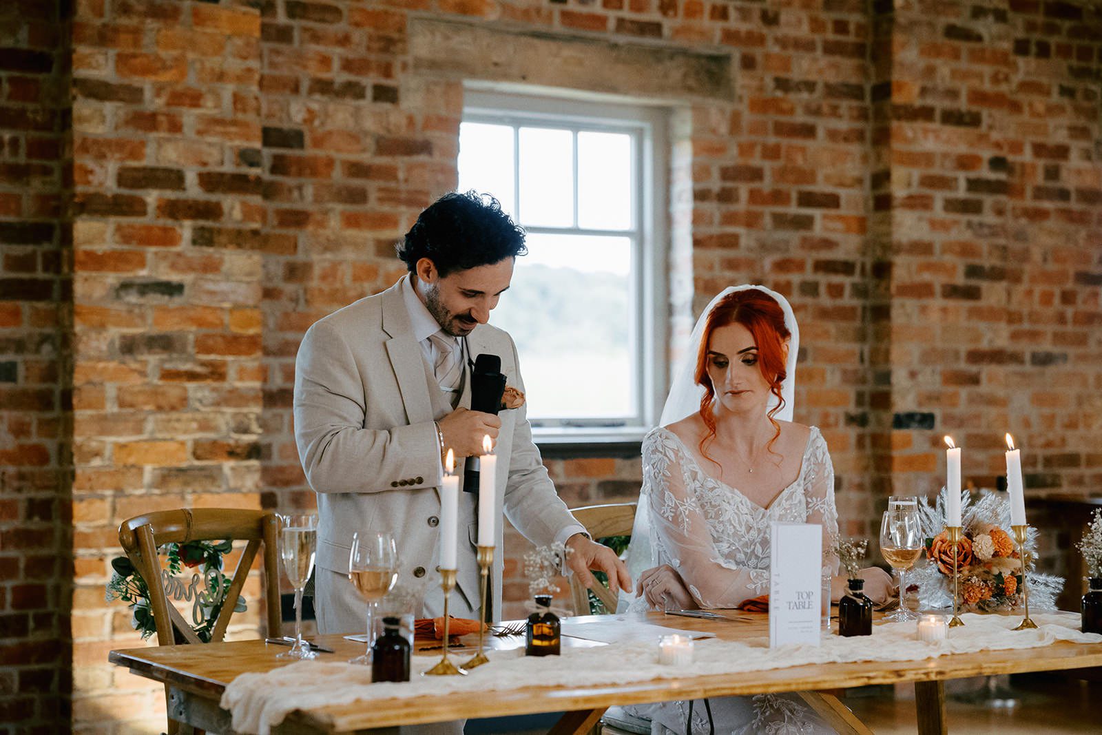 Groom giving his speech at the top table against an exposed brick wall as the bride reads along