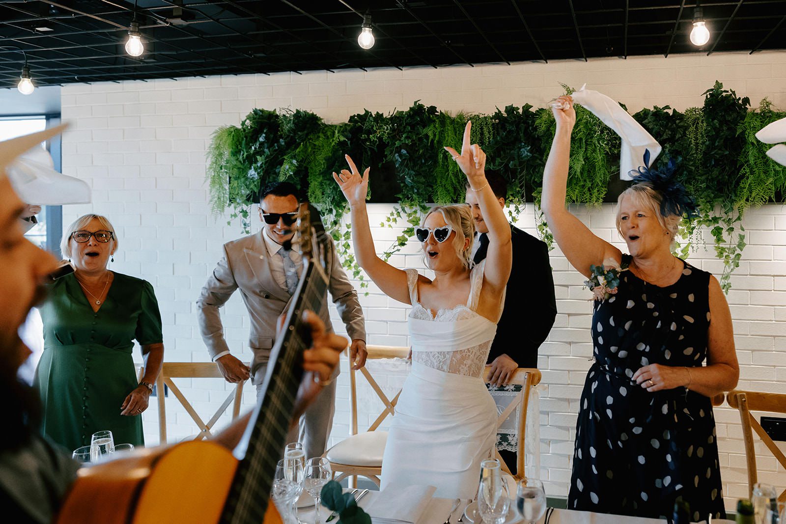 Bride in heart-shaped sunglasses cheering with guests while a guitarist from the day band plays
