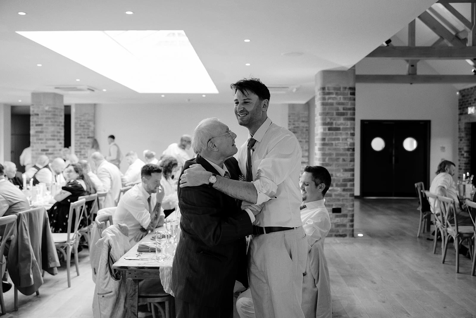 Black and white photo of the groom sharing a big hug with an older guest in the reception barn