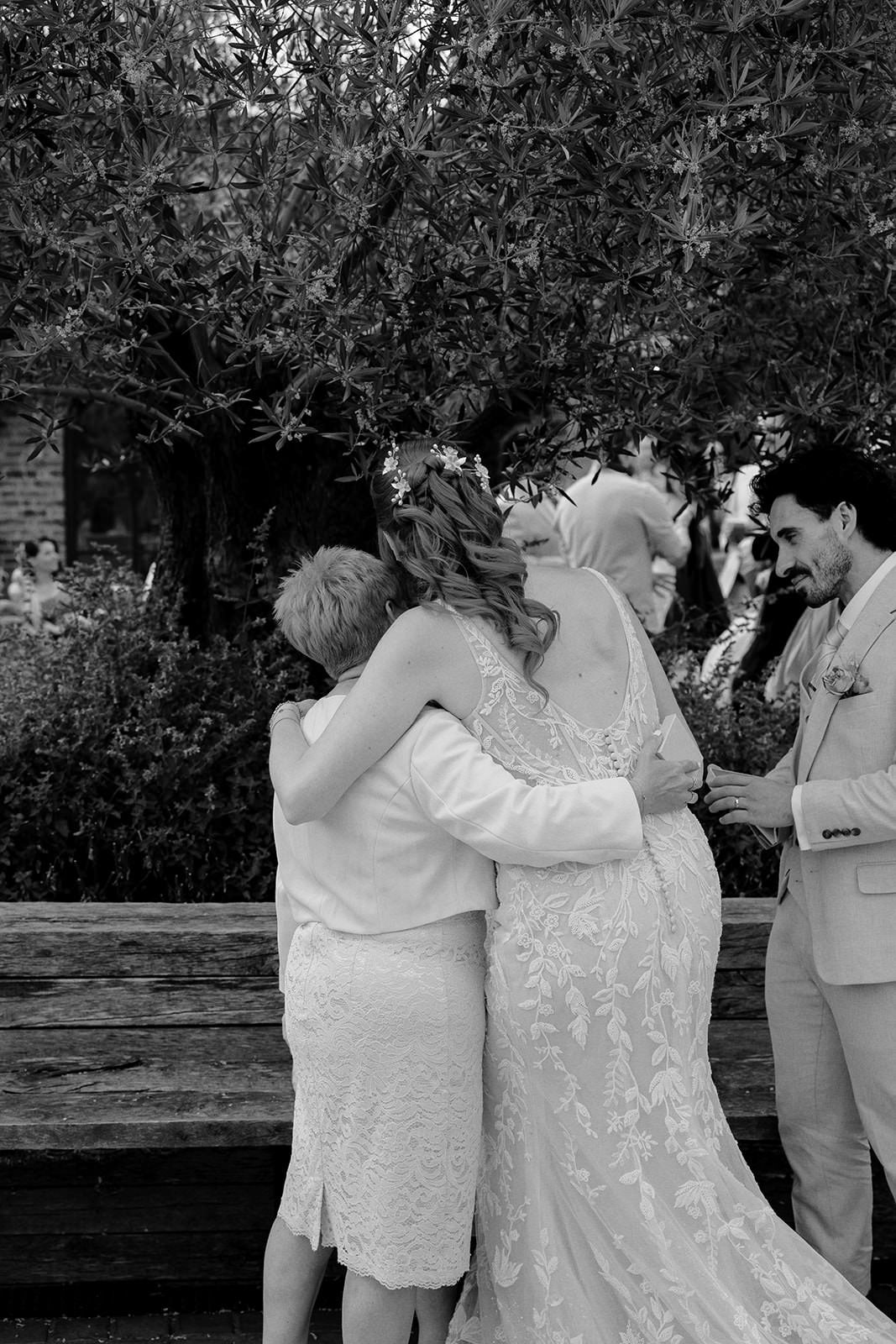 Black and white photo of the bride hugging a guest beneath the courtyard olive tree as the groom looks on