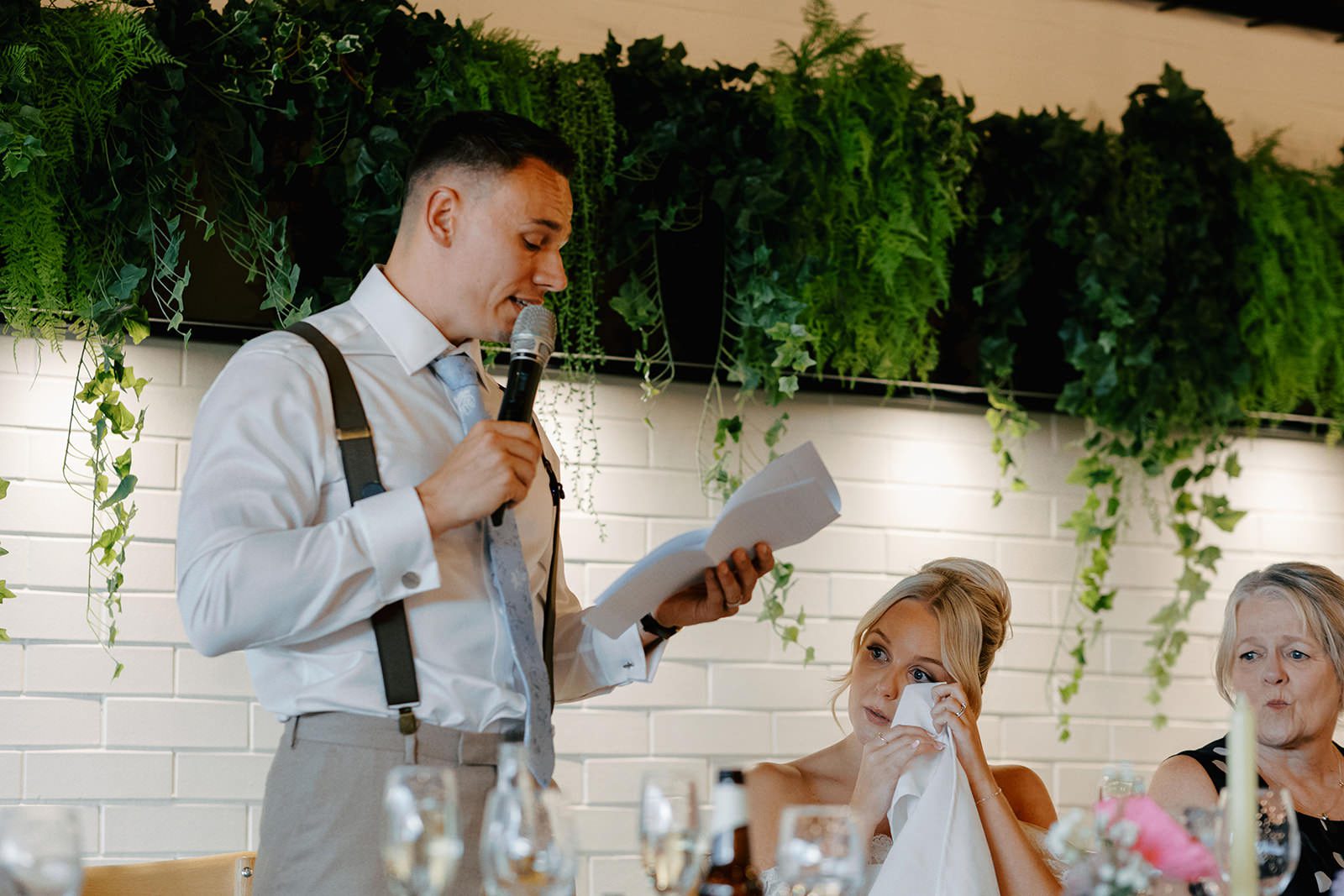 Groom giving his speech in front of a white brick wall with hanging greenery as the bride dabs her eyes