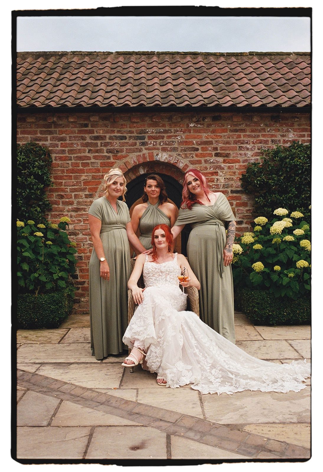 35mm film photo of the bride seated with her bridesmaids in front of a brick archway and hydrangeas