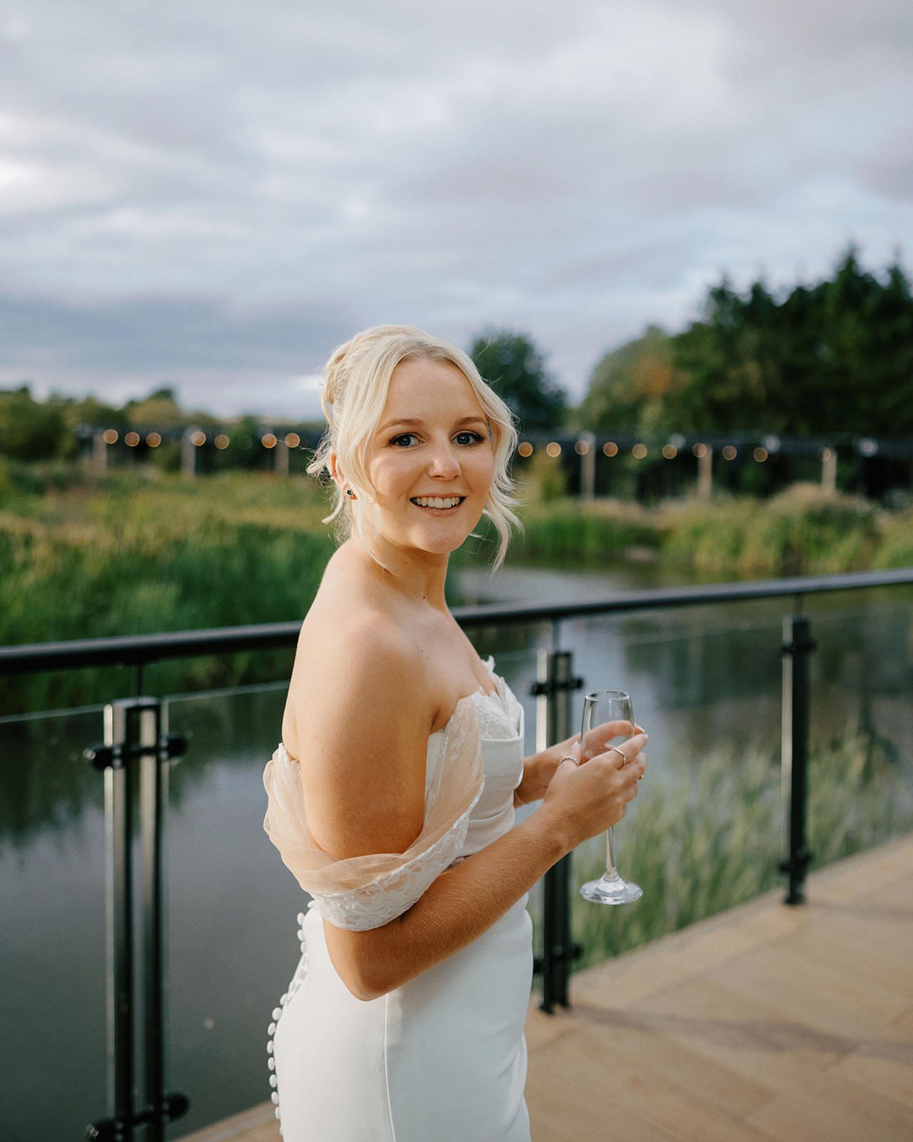 Bride smiling over her shoulder holding a glass of fizz on the lakeside terrace