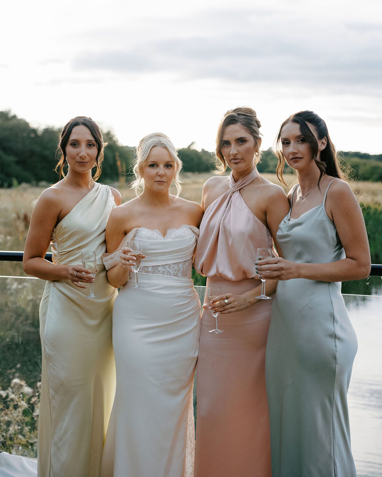 Bride with three bridesmaids in champagne, blush and sage satin dresses beside the lake