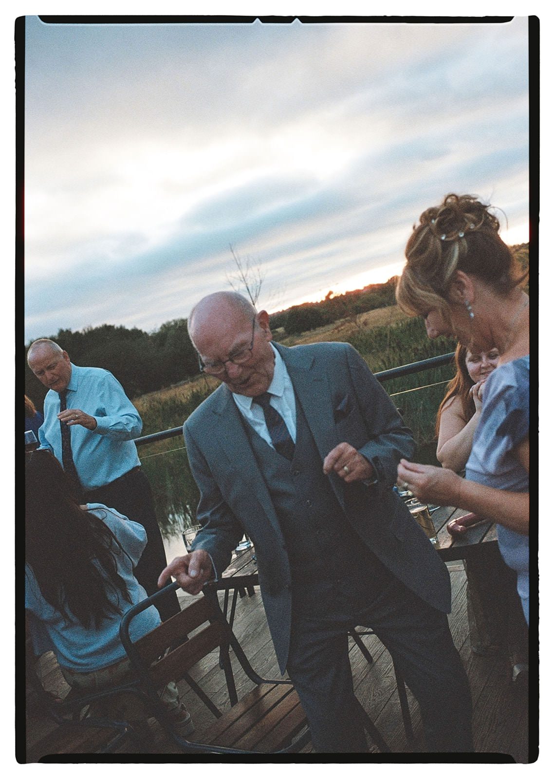 35mm film photo of the bride's grandad laughing with guests on the terrace at dusk