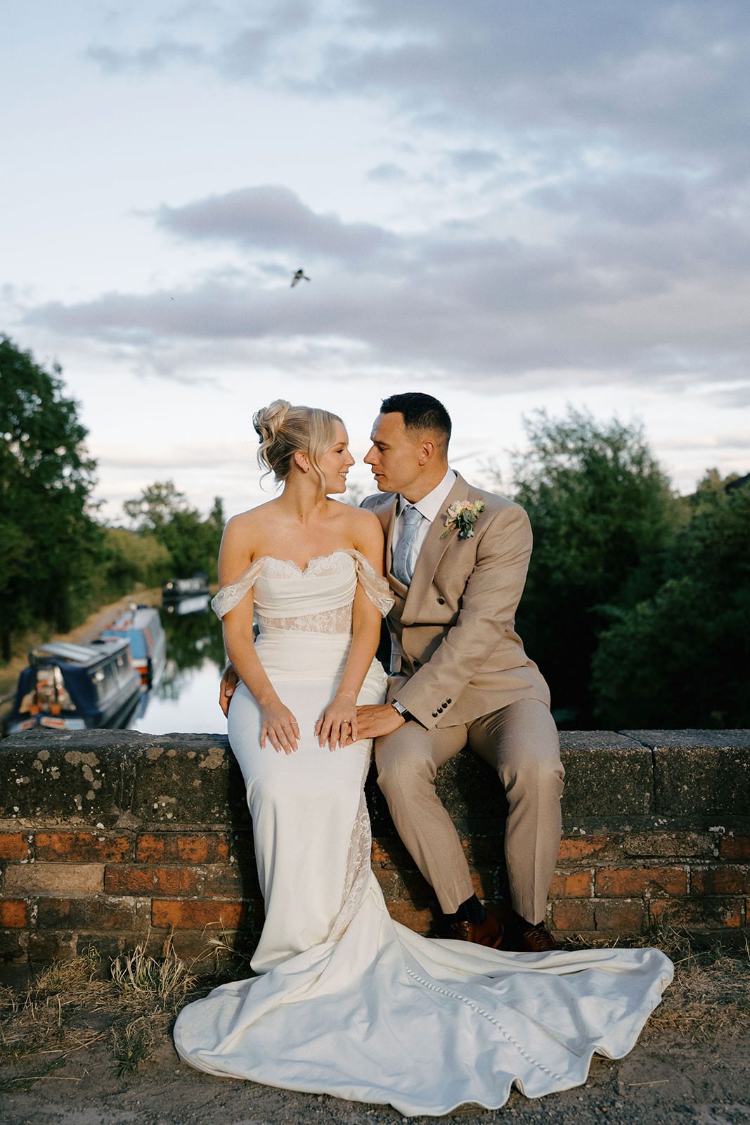 Bride and groom sitting on a brick canal bridge with narrowboats moored behind them