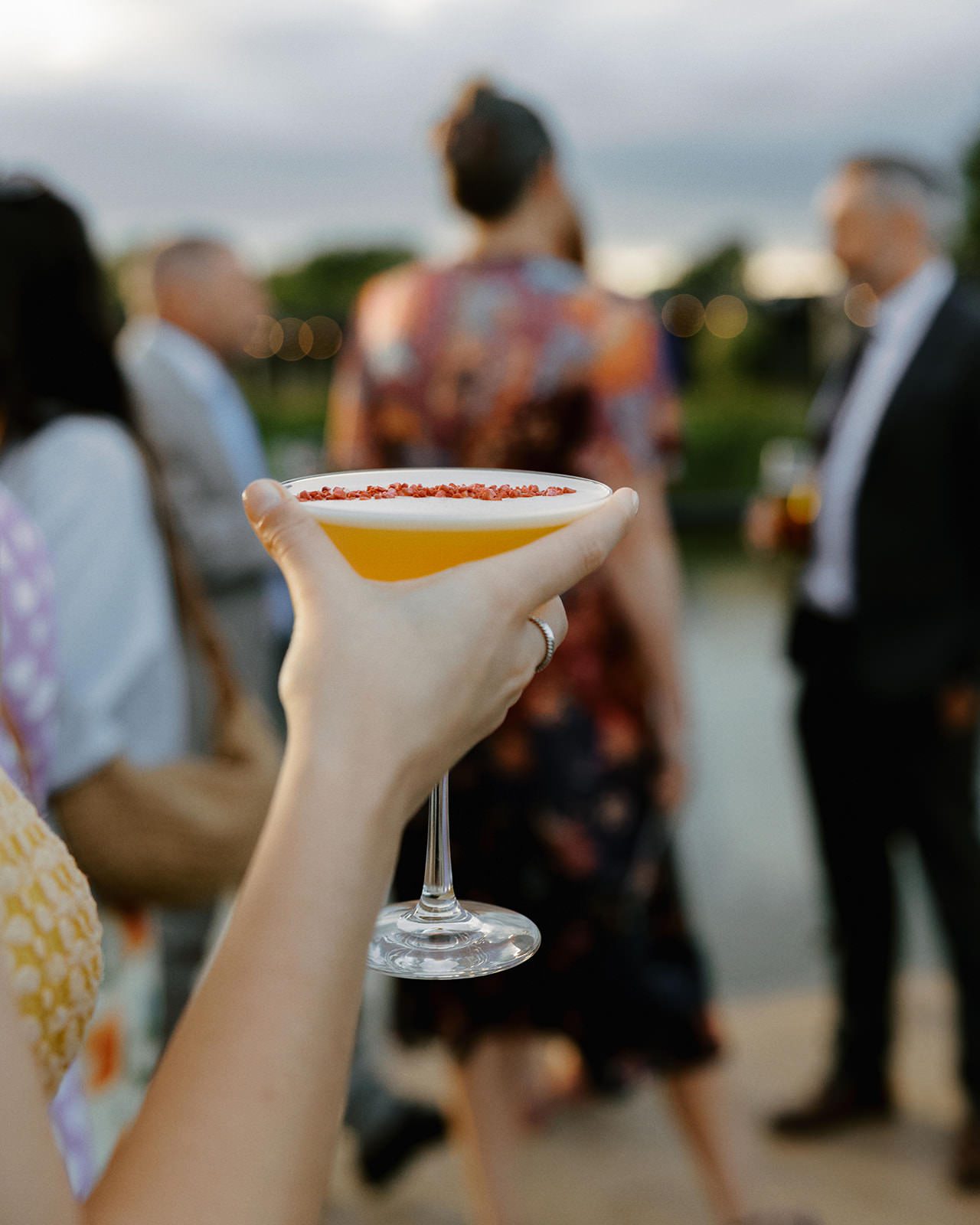 Guest holding a cocktail with guests chatting by the water in the background