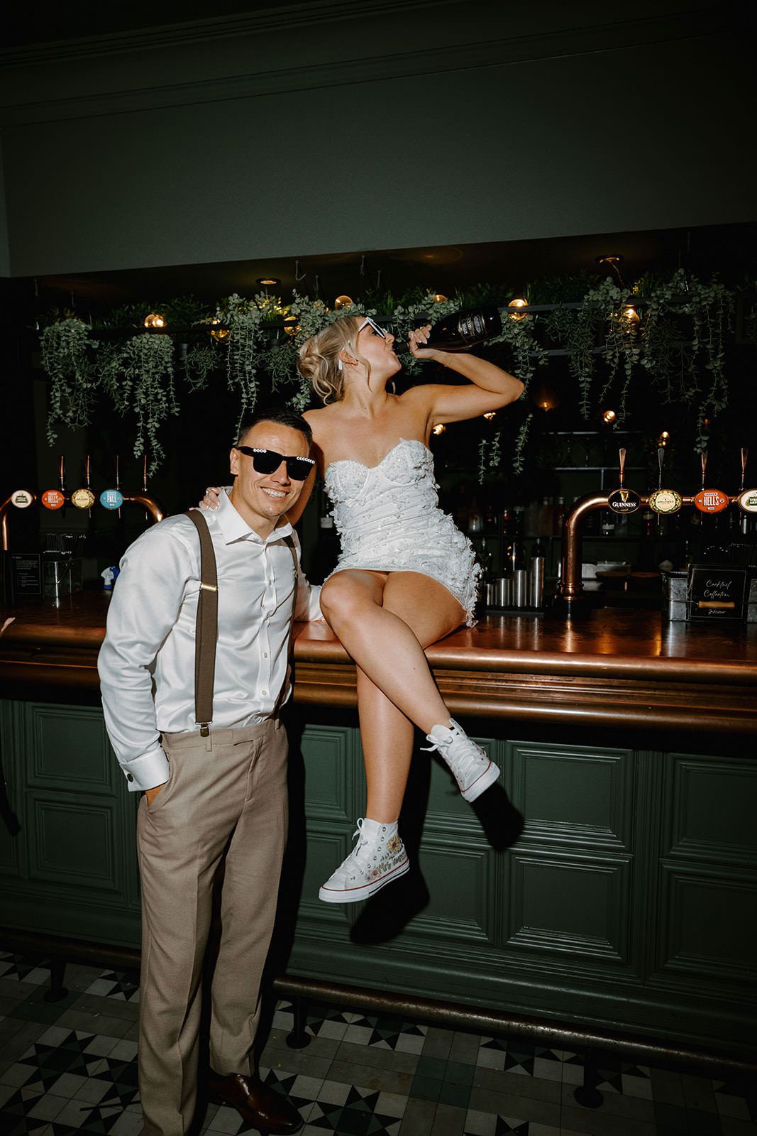 Bride sitting on the bar of the Whistle Barns private pub with the groom in sunglasses beside her