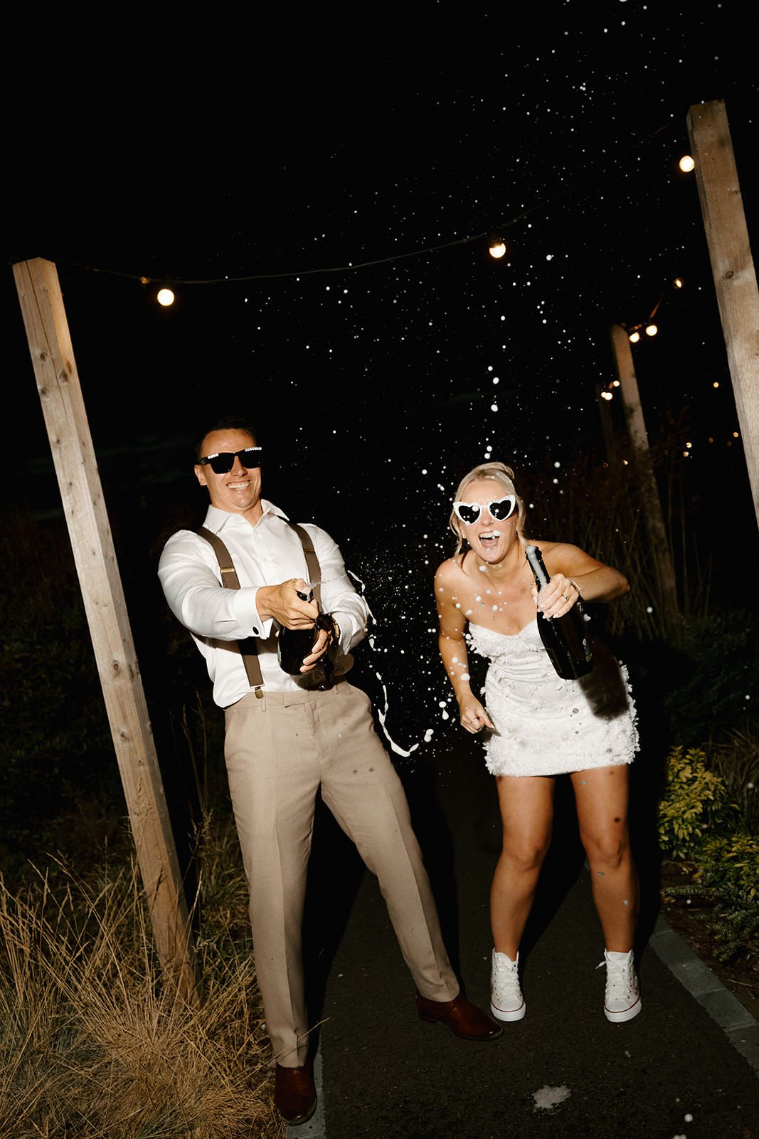 Bride and groom in sunglasses spraying champagne under festoon lights at night