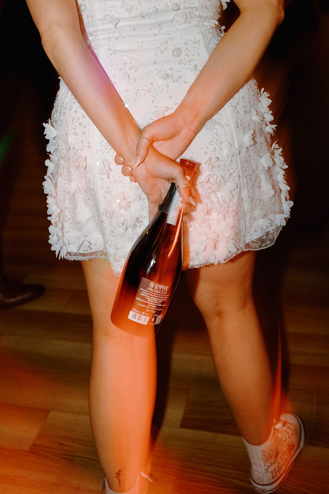Detail of the bride's 3D floral mini dress and Converse trainers, holding a bottle behind her back